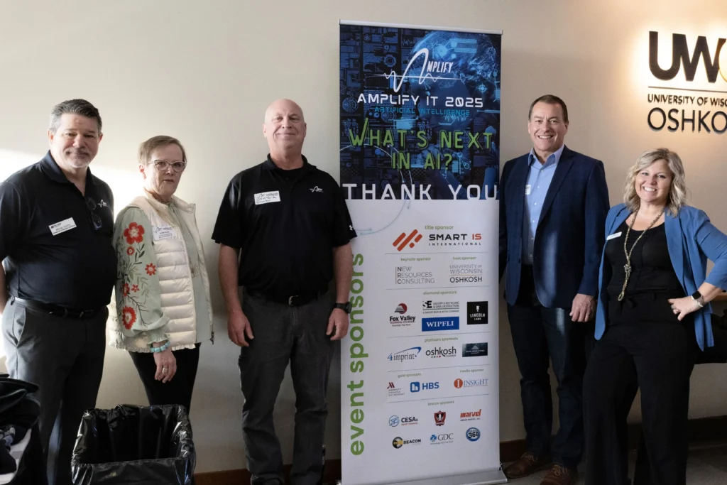 Five professionally dressed individuals standing beside an event sponsor banner for "Amplify IT 2025" at University of Wisconsin Oshkosh