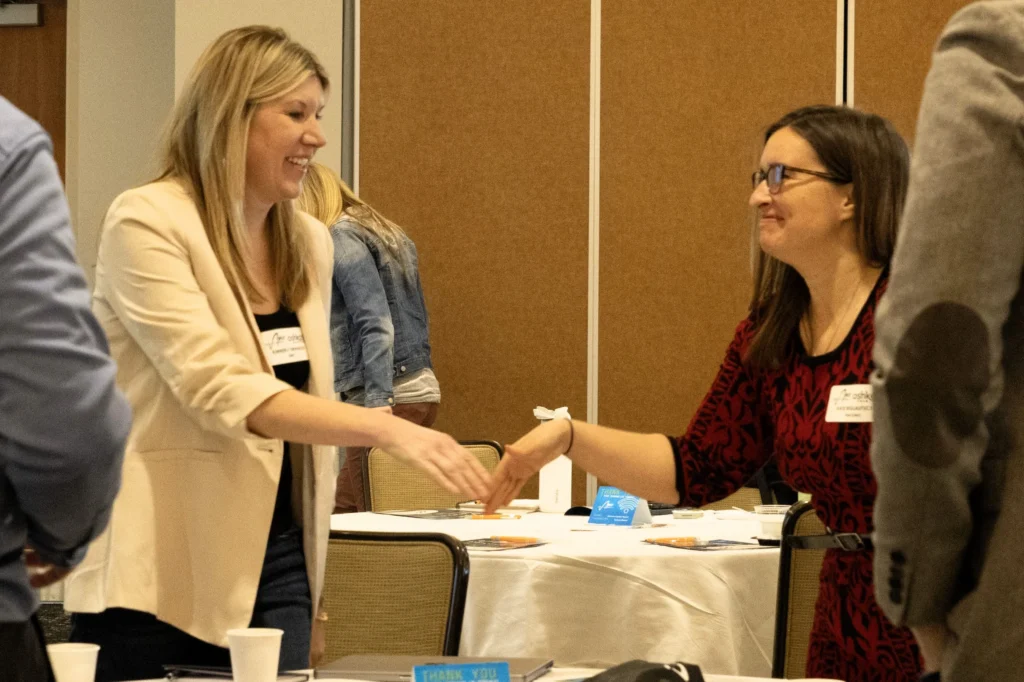 Two women smiling and shaking hands in a professional conference setting.