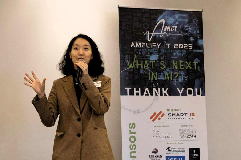 Female speaker in brown blazer presenting near "Amplify IT 2025" AI conference banner.