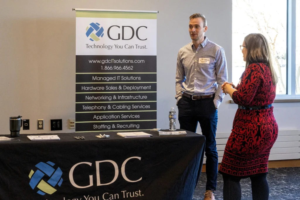 Two professionals conversing at a GDC IT solutions booth with informational banner and table.