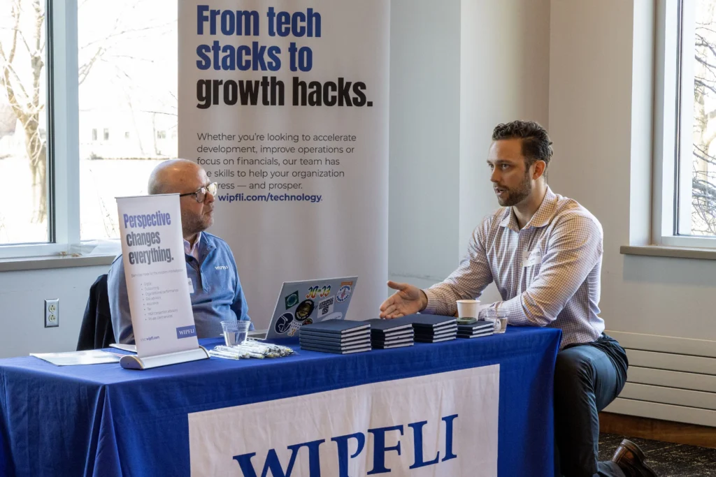Two men seated at a Wipfli company table with promotional banners and materials in a professional setting.