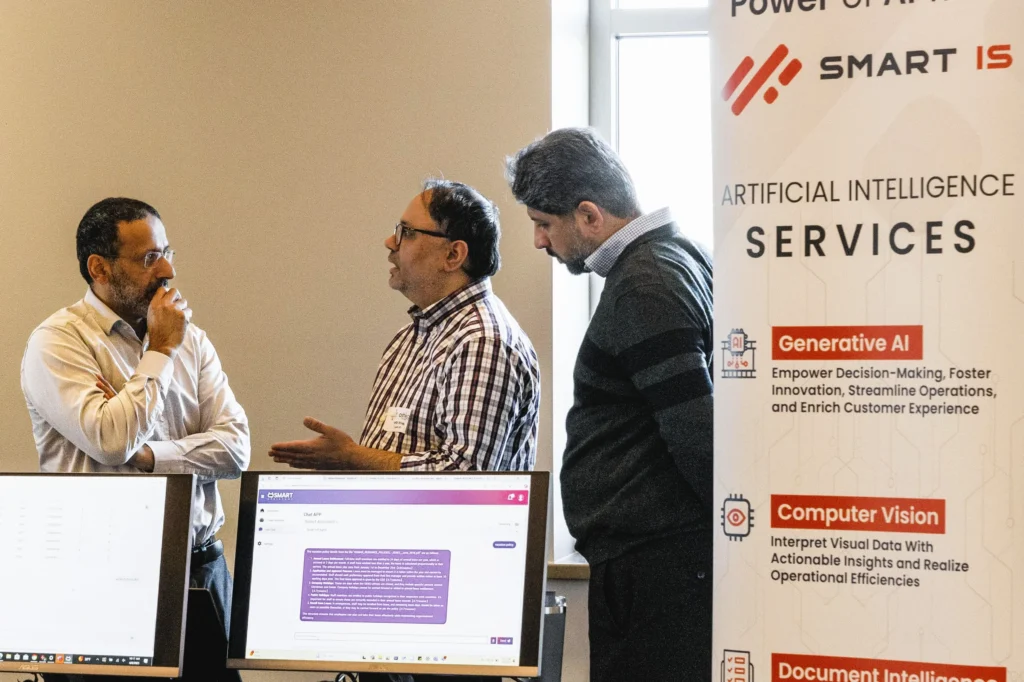Three men engaged in discussion beside computer monitors and a banner detailing AI services.