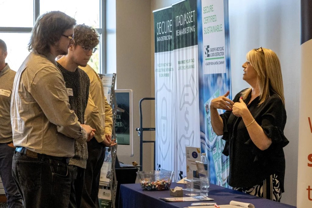Professional woman explaining services at trade show booth to two male attendees.