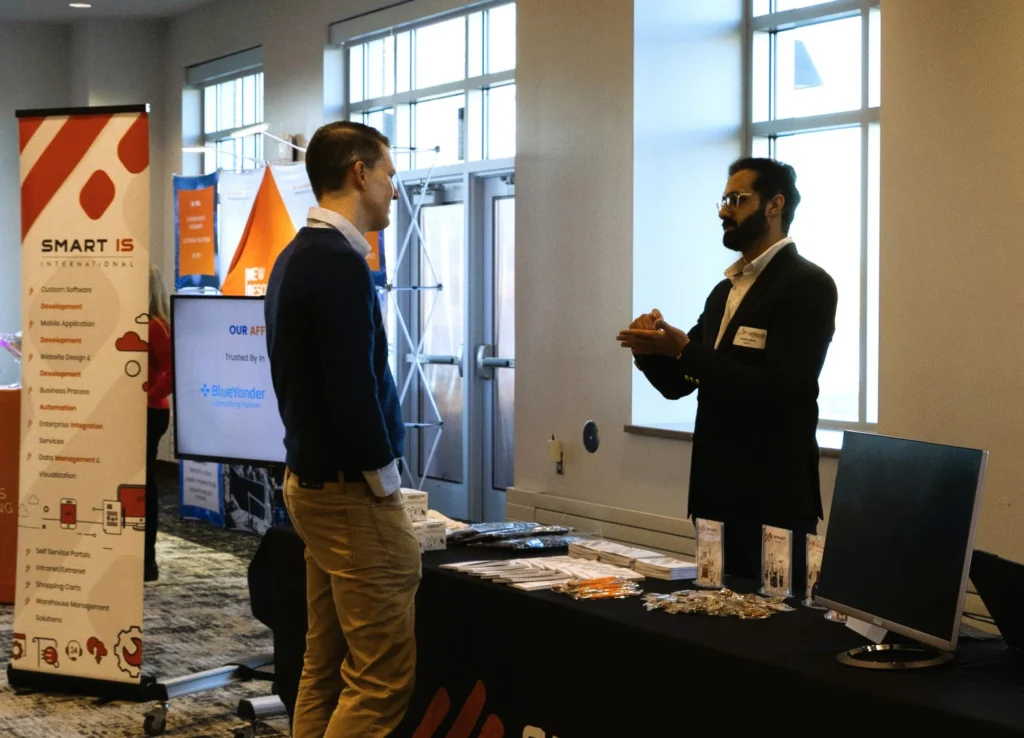 Two men engaged in conversation at a Smart IS International promotional booth with brochures and merchandise.