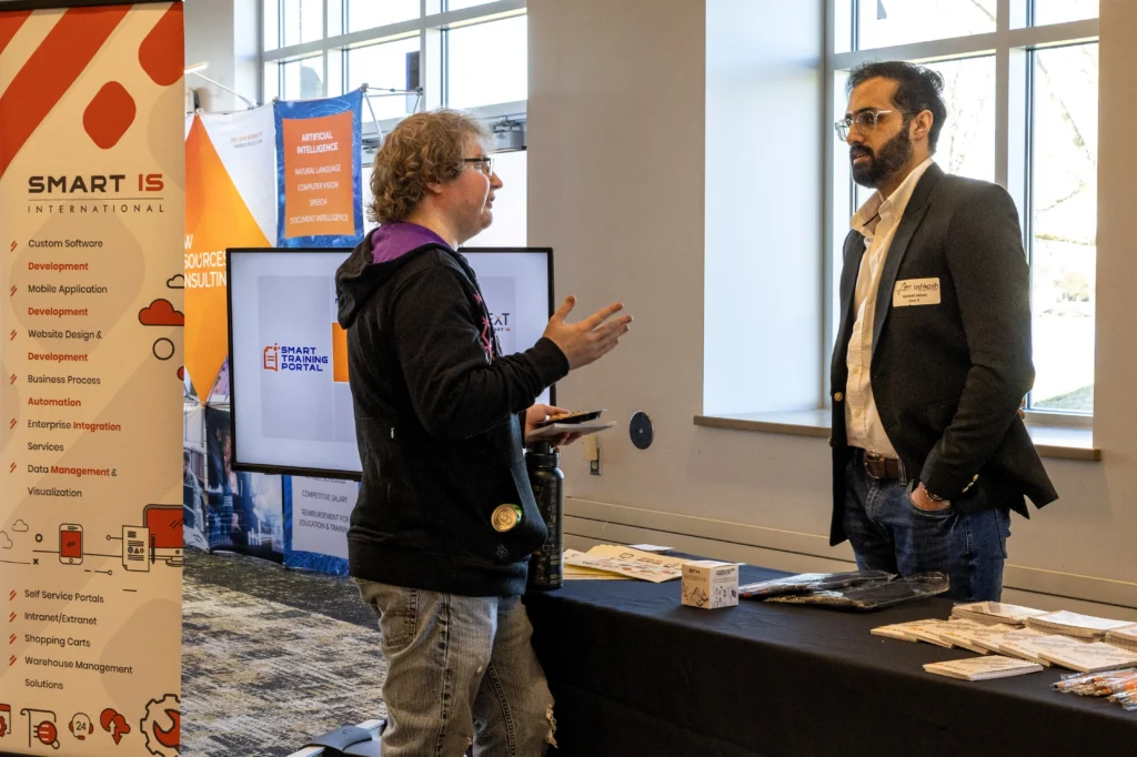 Two men conversing at a professional event booth with promotional banners and materials.