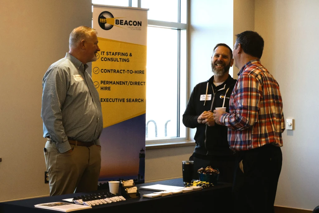 Three men conversing at a Beacon IT staffing and consulting promotional booth with a yellow and blue banner.