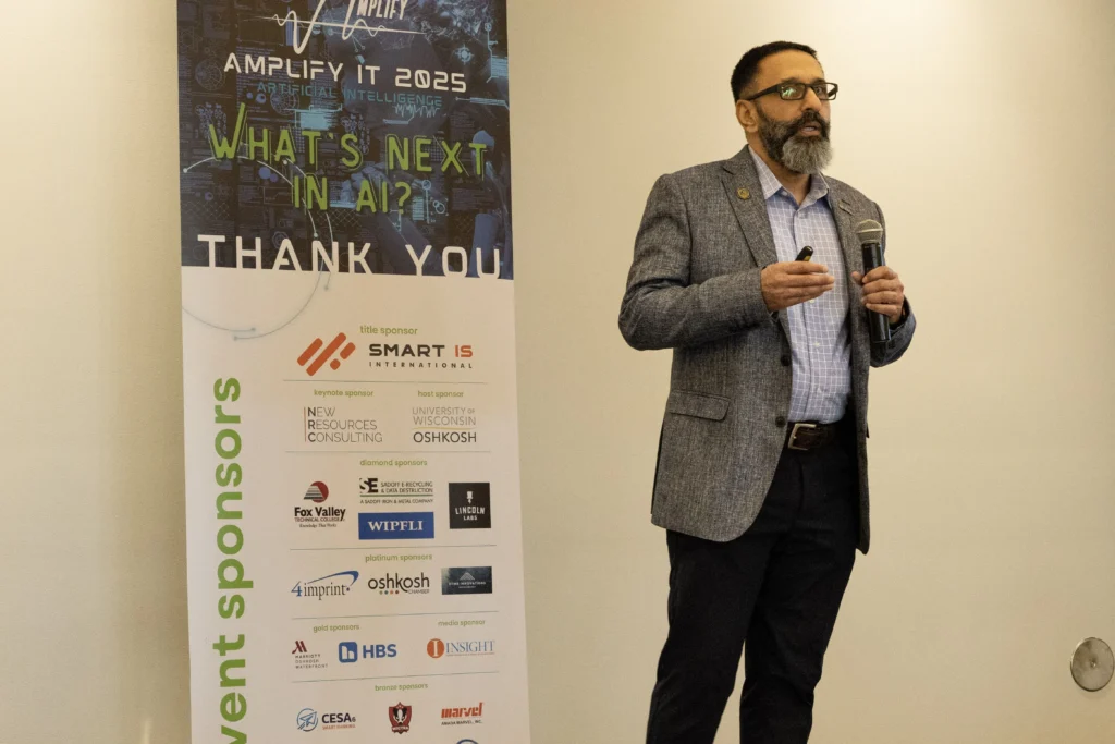 Professional man with beard and glasses speaking at AI conference next to sponsor banner.