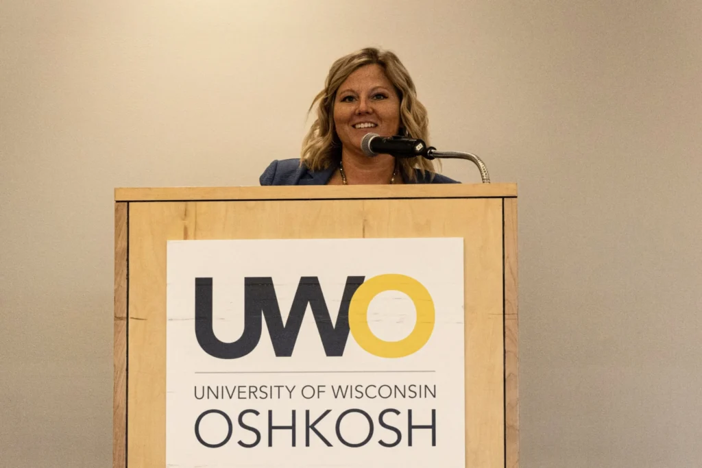 Woman speaking at a wooden podium with University of Wisconsin Oshkosh logo.