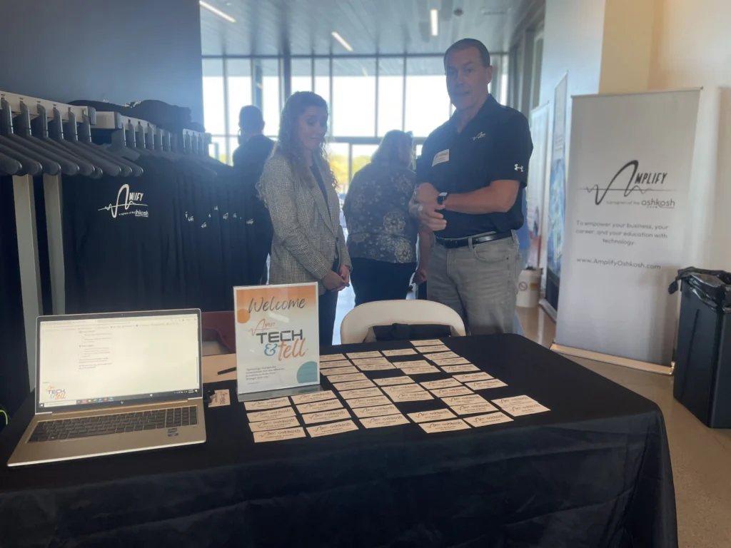 Professional event booth with two representatives, name tags on table, black Amplify Oshkosh shirts, and informational signage.