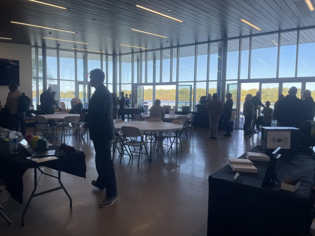 Interior view of a spacious event room with large windows overlooking water, people standing and sitting around tables.