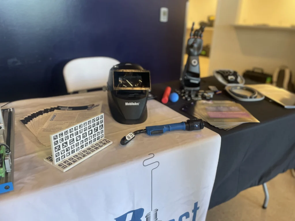 Exhibition table with welding helmet, robotic hand, blue tool, and informational brochures on white and black tablecloths.