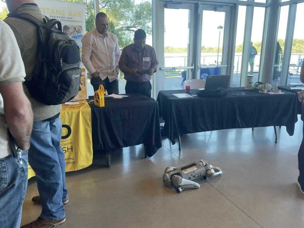 Exhibition booth with two men standing behind tables displaying technology and a four-legged robot on the floor.