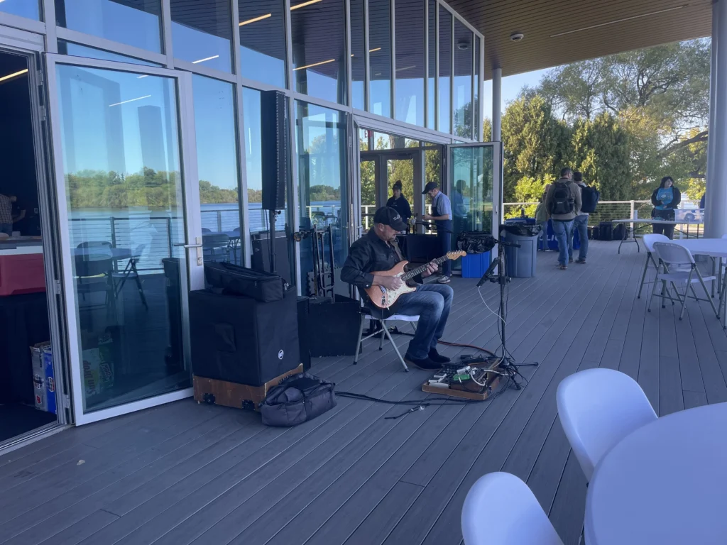 Man playing electric guitar on outdoor patio near glass building with tables and people.