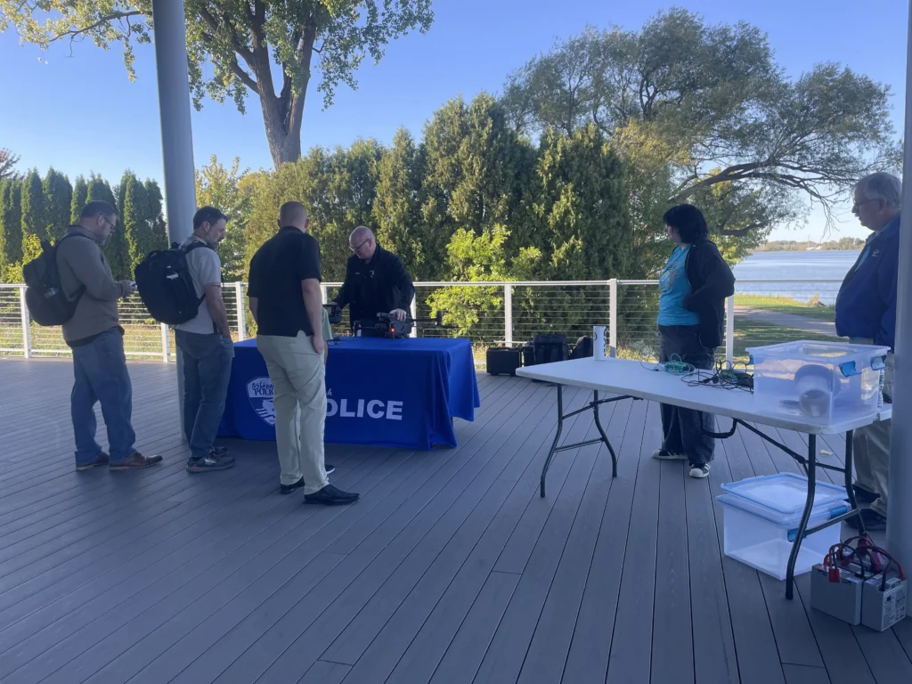 Group of people gathered around a police table with drone equipment on an outdoor deck near water.