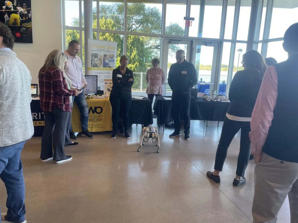 Group of people observing a small robotic dog demonstration near University of Wisconsin Oshkosh engineering display.