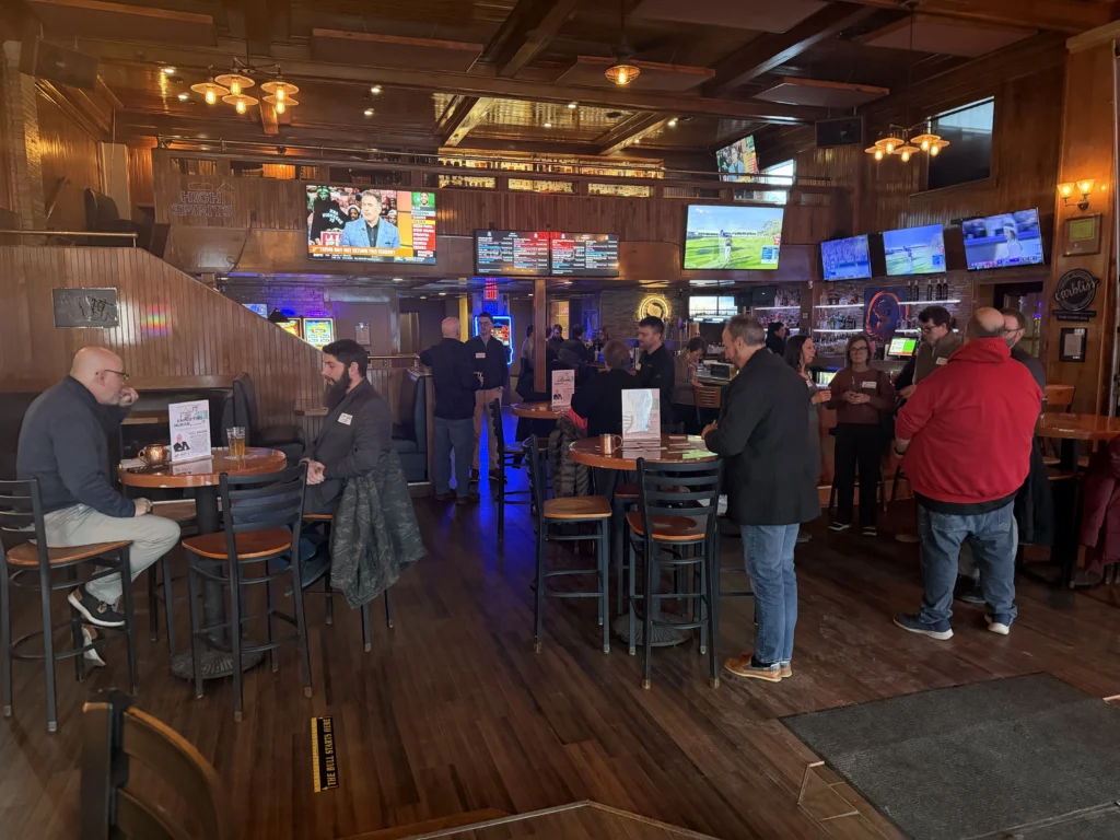 Interior view of a wood-paneled bar with patrons seated and standing, multiple televisions displaying sports and news.