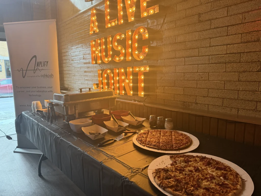 Buffet table with pizzas, condiments, and serving dishes under illuminated "Alive Music Joint" sign.