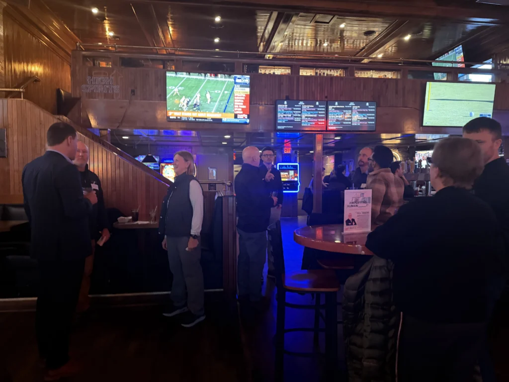 Interior of a dimly lit bar with people socializing near wooden tables and multiple TVs displaying sports.