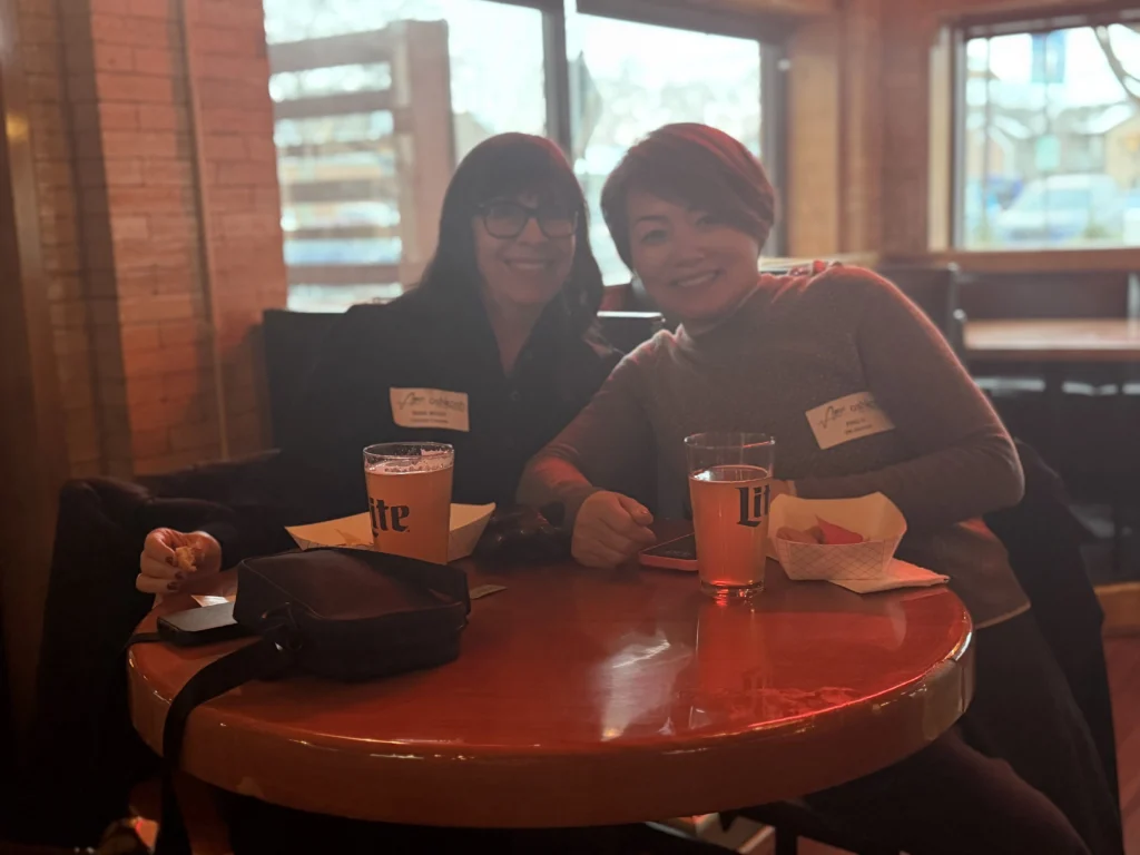 Two women with name tags seated at a wooden table with drinks and snacks in a dimly lit restaurant.