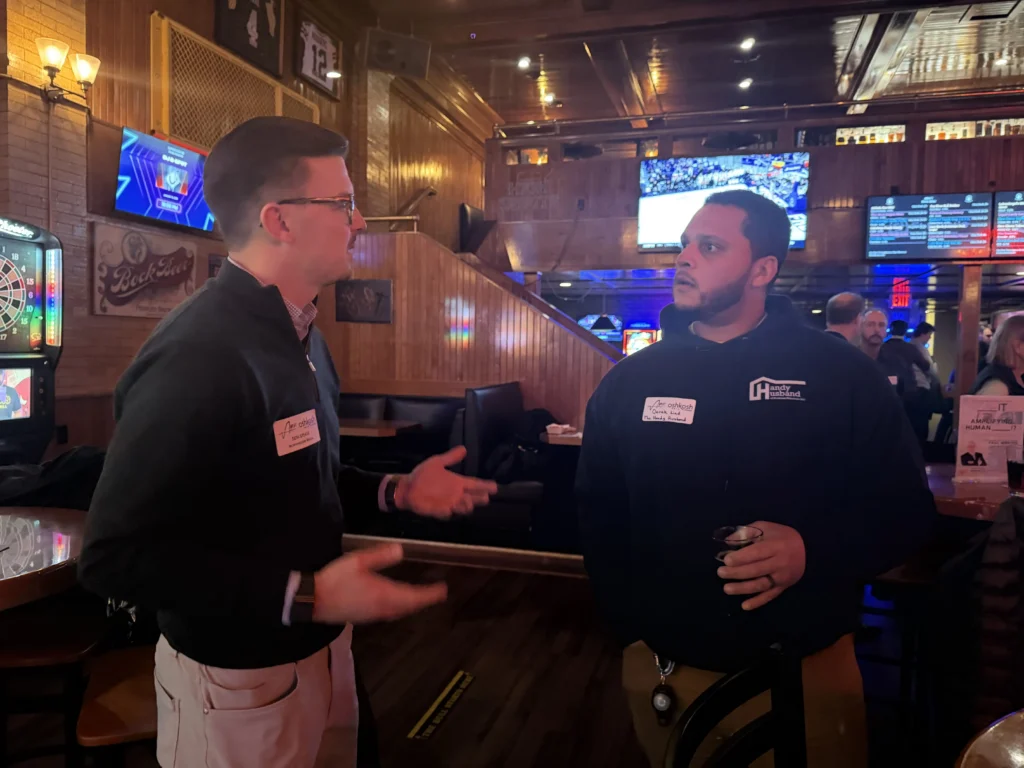 Two men wearing name tags engaged in conversation inside a dimly lit bar with wooden interior.