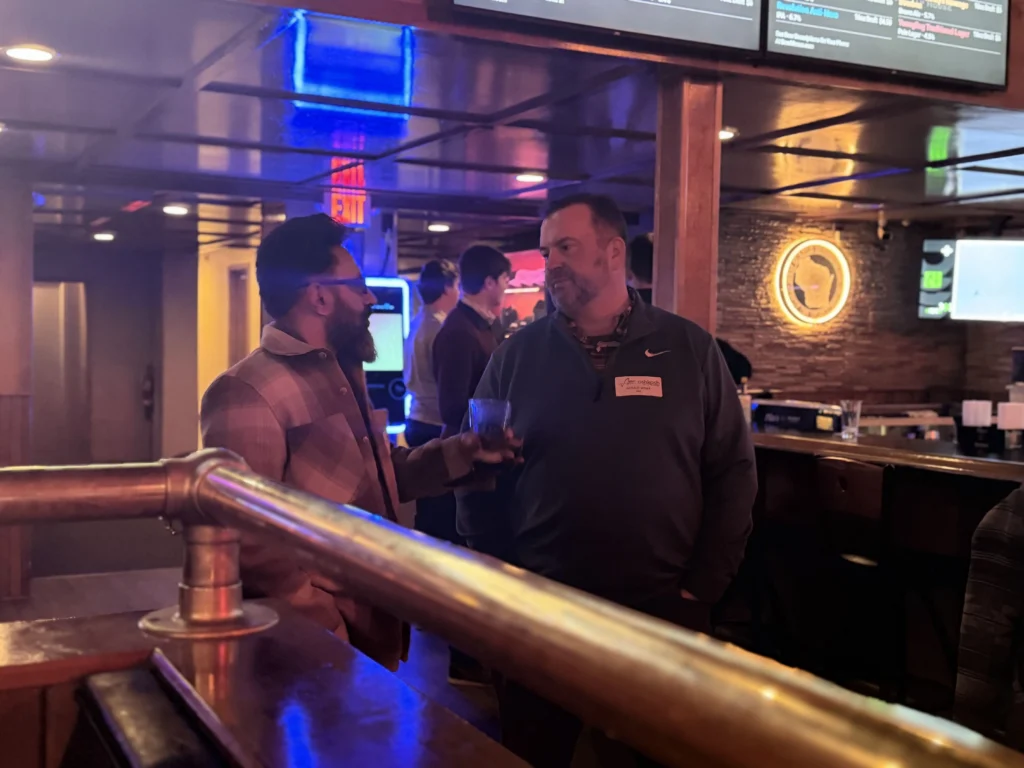 Two men conversing in a dimly lit bar with neon signs and a digital menu overhead.