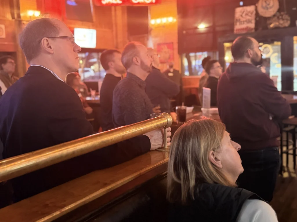 Group of adults attentively watching an event inside a warmly lit bar or restaurant.