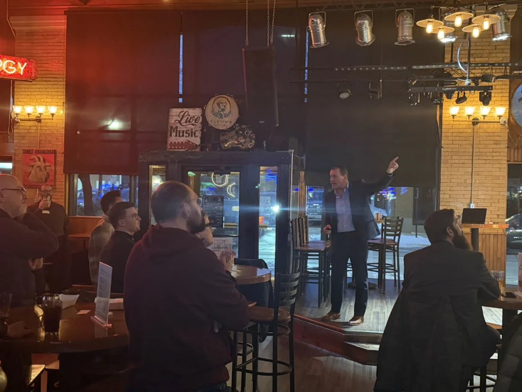 Man in formal attire speaking to attentive audience in dimly lit bar with "Live Music" sign.