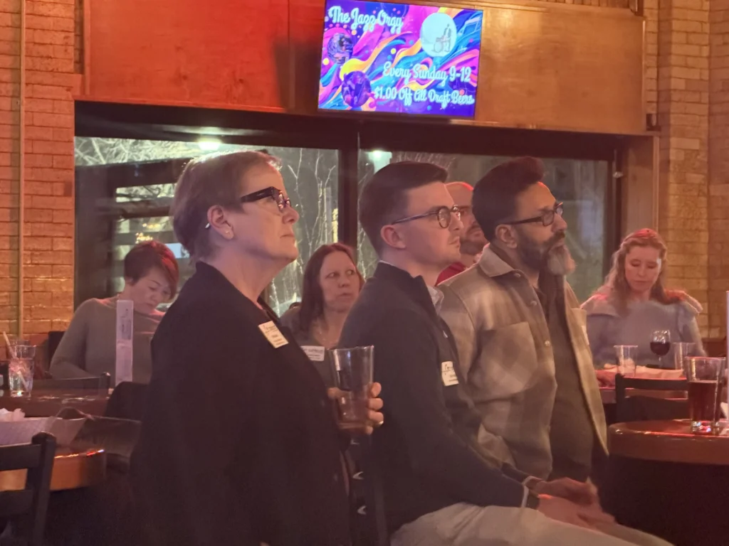 Group of diverse adults seated indoors, attentively listening, with colorful "The Jazz Orgy" sign overhead.