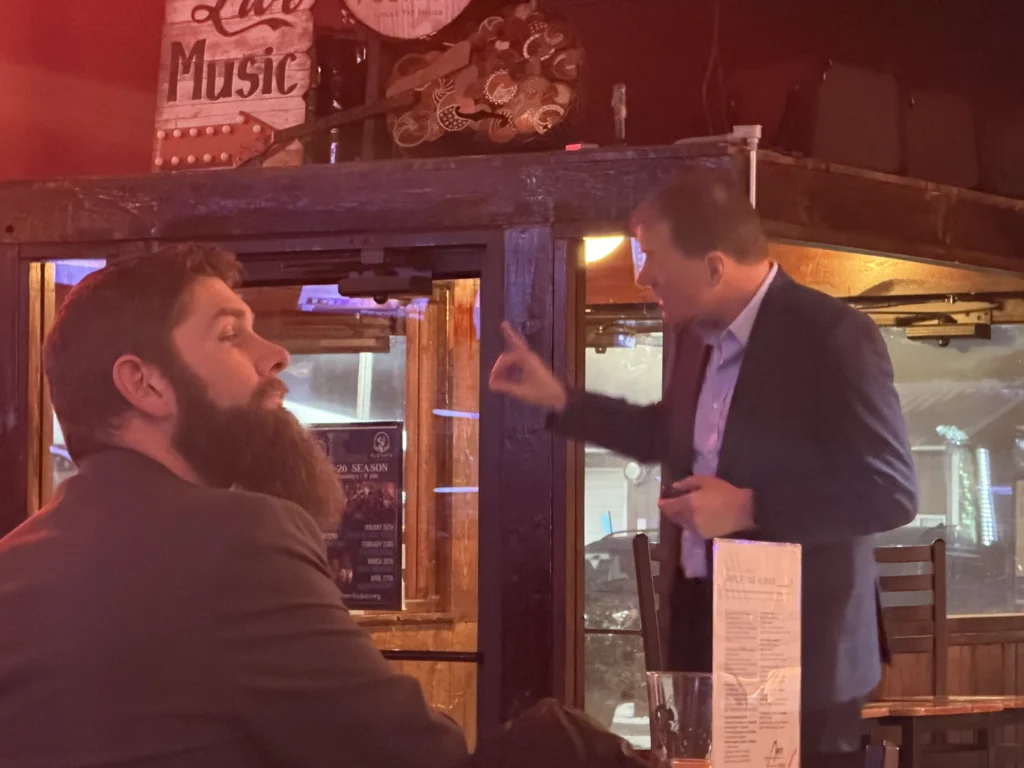 Two men in a dimly lit bar with a "Live Music" sign and a menu on the table.