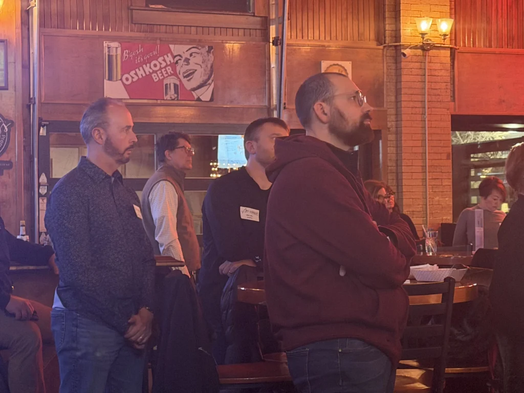 Group of men attentively standing in a dimly lit bar with wooden paneling and vintage beer signage.