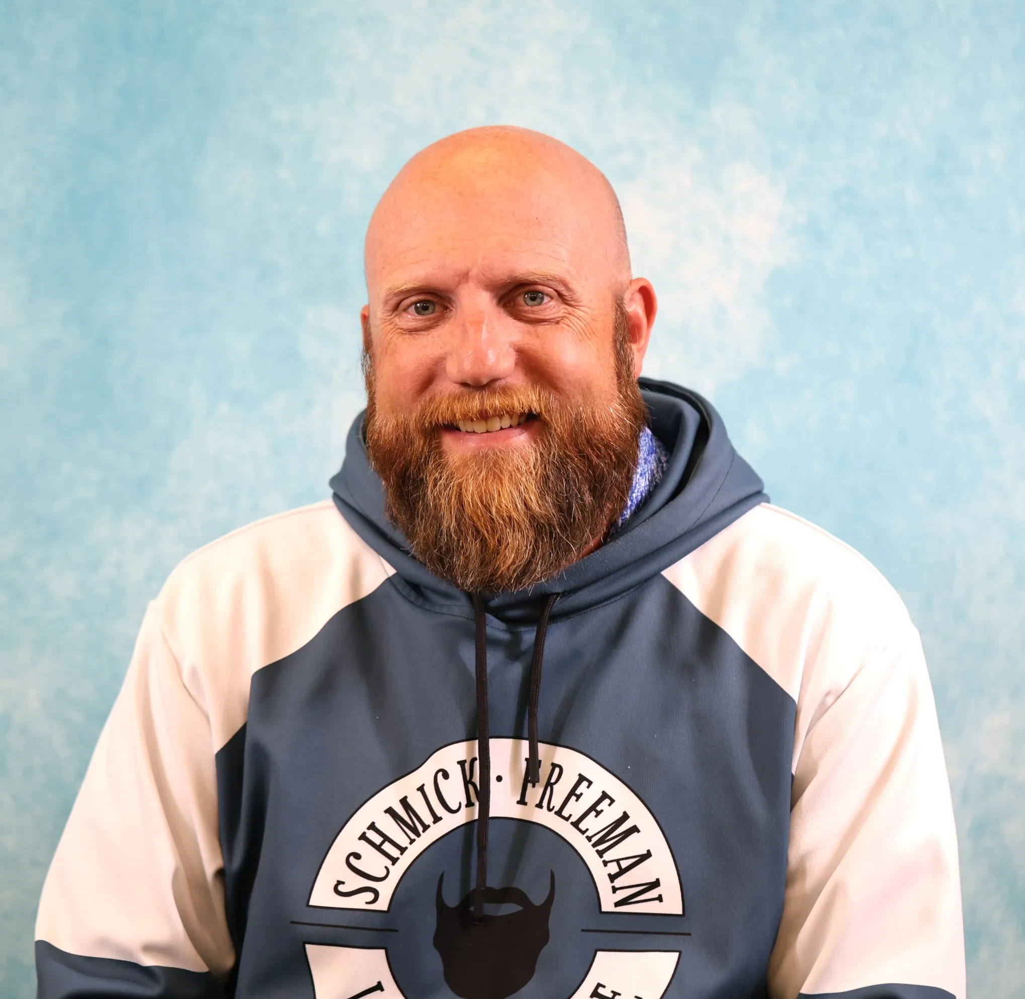 Portrait of a smiling bald man with a red beard wearing a navy and white hoodie.
