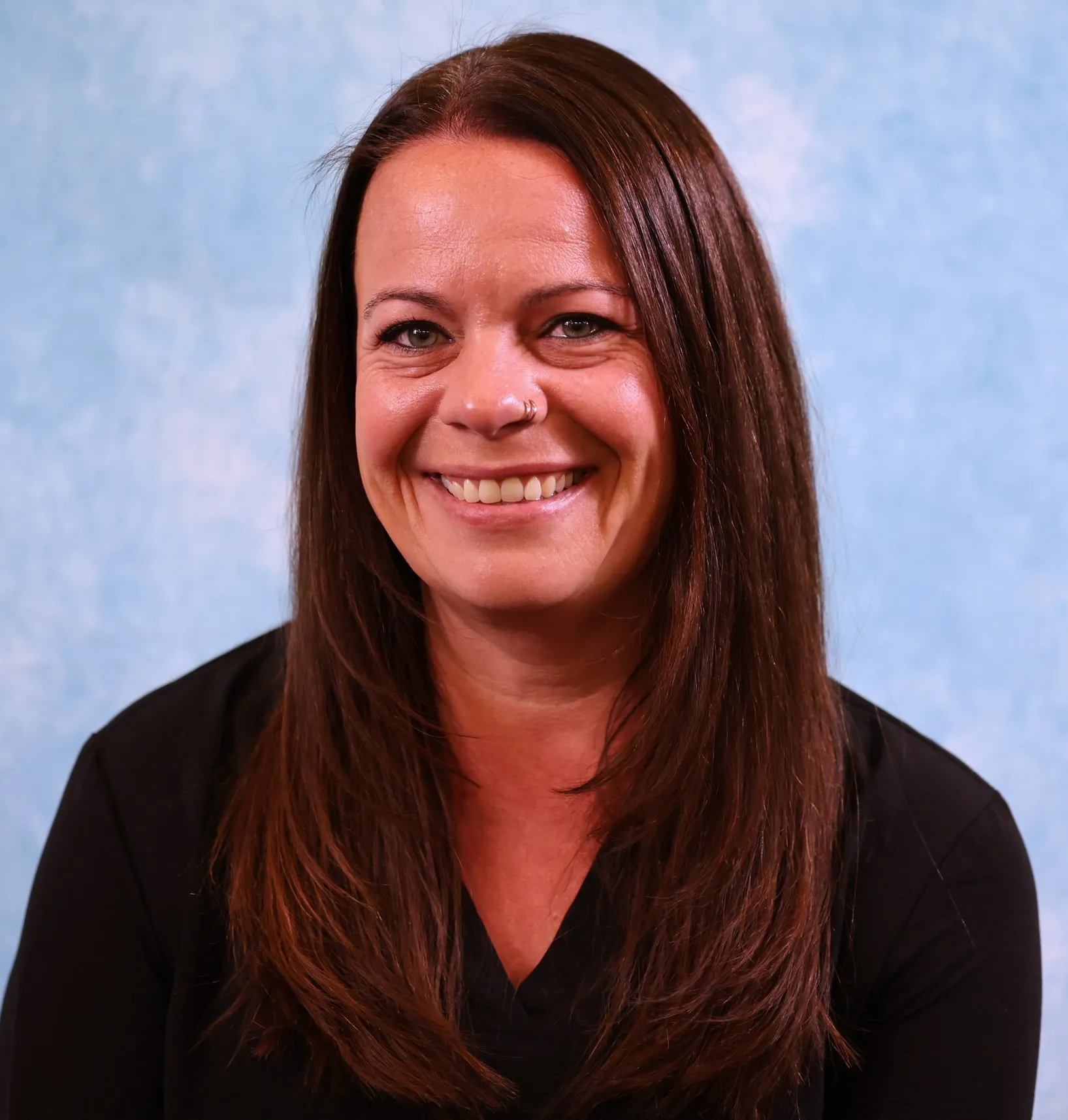 Portrait of a smiling woman with long brown hair wearing a black top against a blue background.