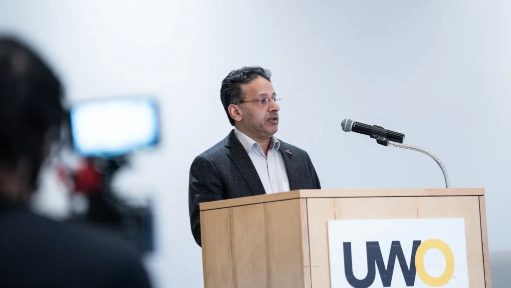 Man in a suit speaking at a wooden podium with a UWO logo and microphone.