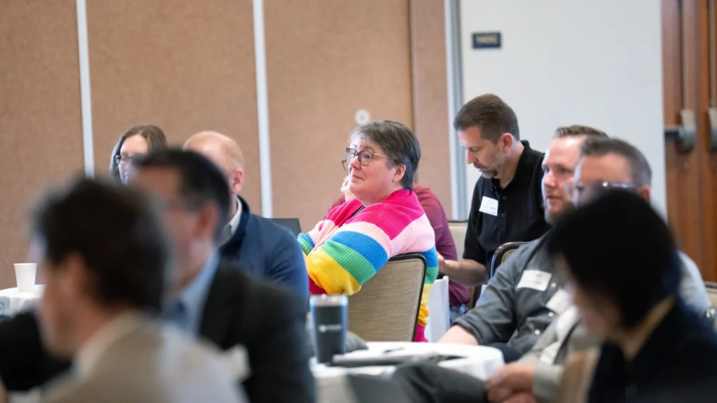Audience of diverse adults seated and attentively listening in a conference room setting.