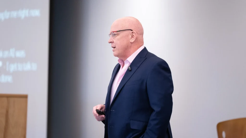 Professional bald man in glasses and navy suit presenting in front of a blurred projection screen.