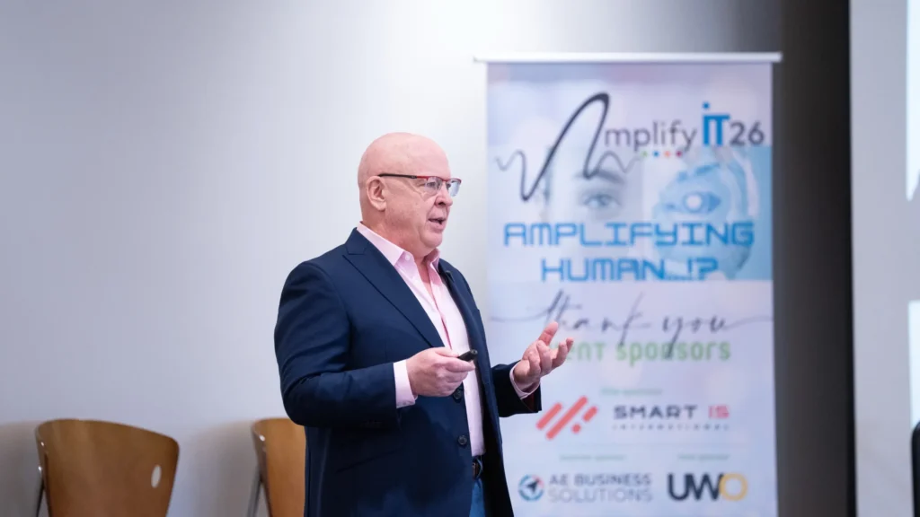 Middle-aged man in a navy suit and pink shirt speaking at Amplify IT 26 conference.