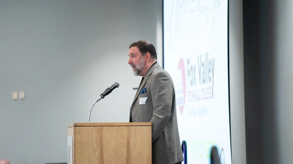 Man in gray blazer speaking at wooden podium with microphone and presentation screen.