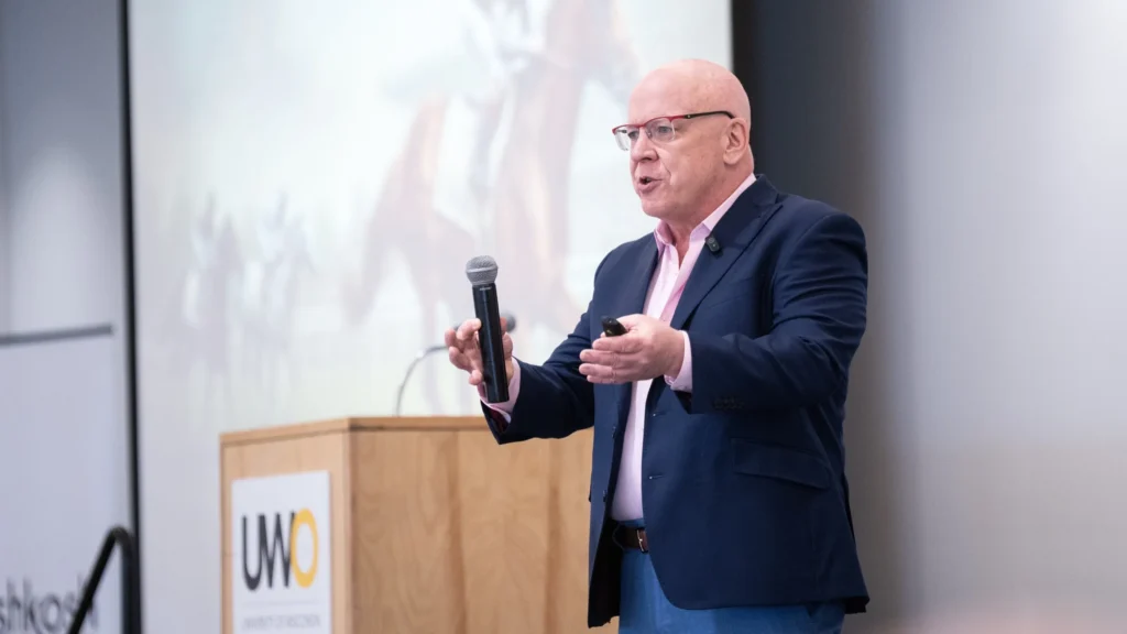 Bald man wearing glasses and a navy blazer speaking with a microphone at University of Wisconsin presentation.