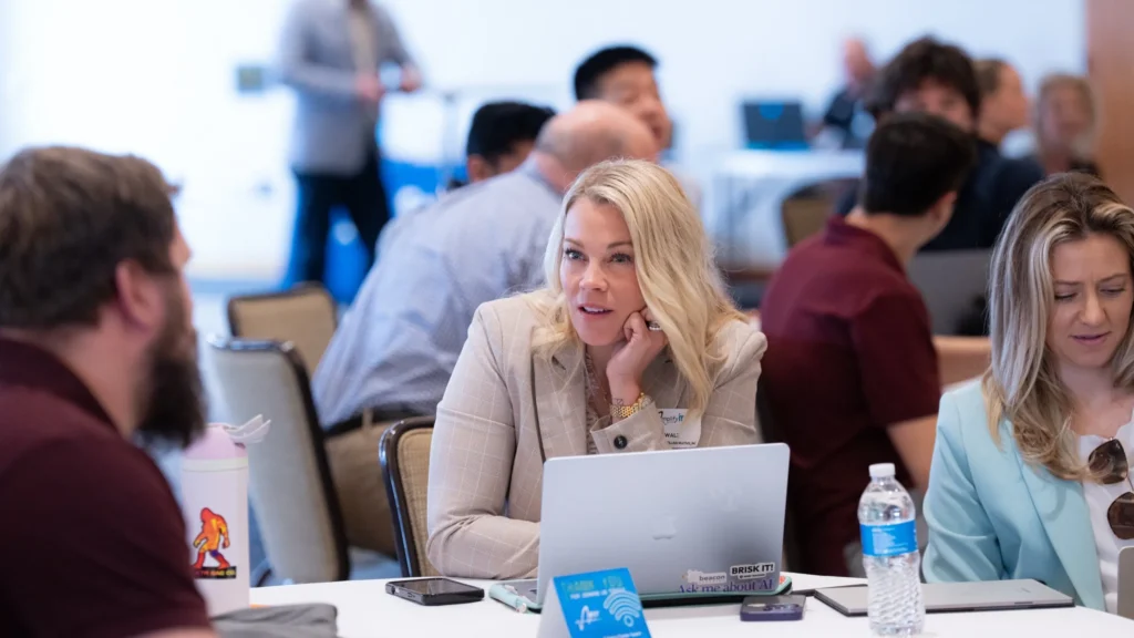 Professionally dressed individuals engaged in discussion around a table with laptops and water bottles.