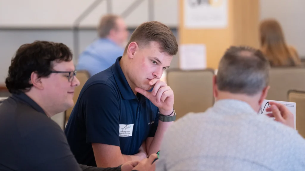 Three men engaged in discussion at a professional meeting, one wearing a blue shirt with a name tag.
