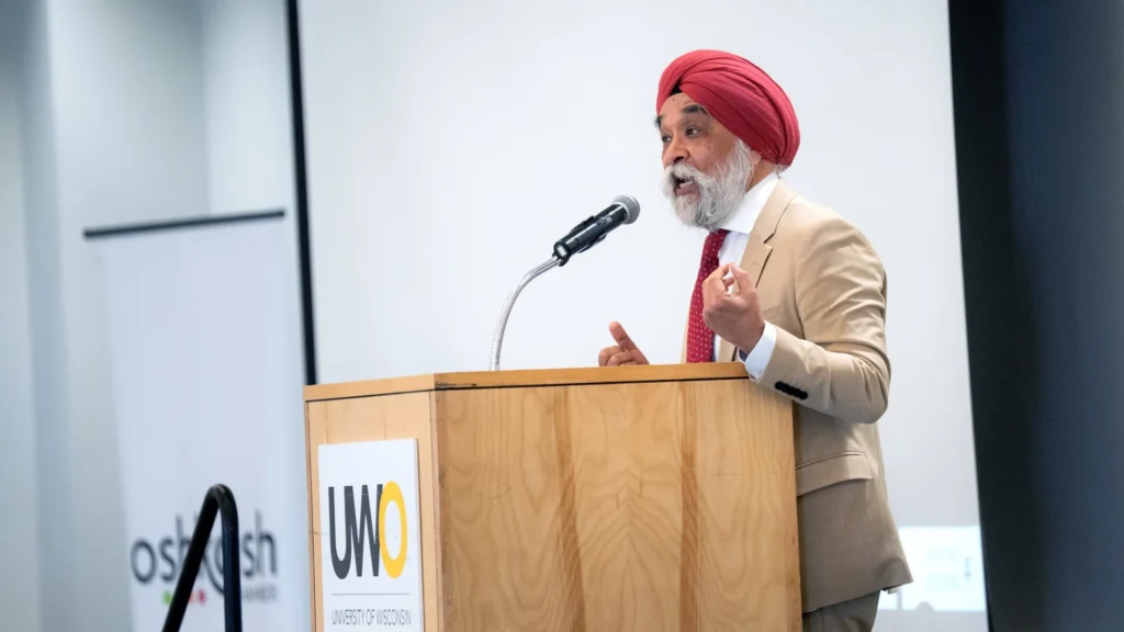 Man wearing a red turban and beige suit speaking at a podium with UW Oshkosh logo.