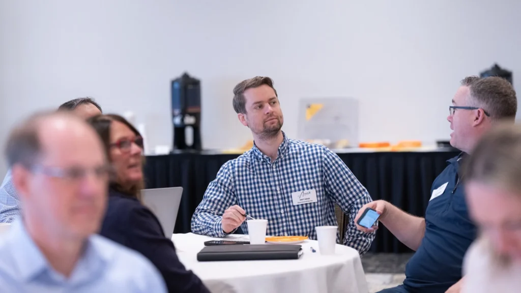 Group of professionals engaged in discussion at a round table with coffee cups and name tags.