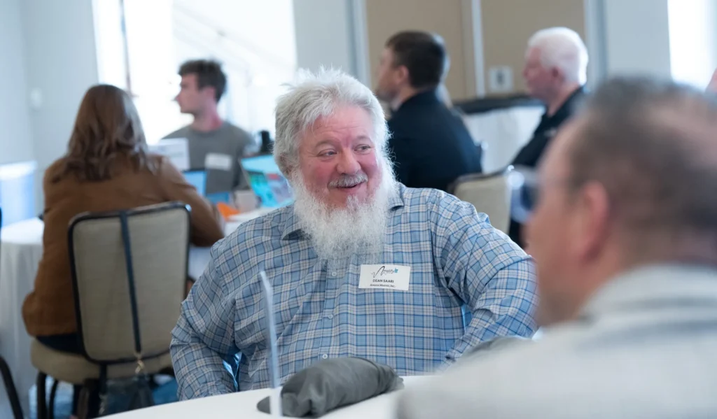 Older man with white beard and plaid shirt wearing a name tag, seated at a conference table.