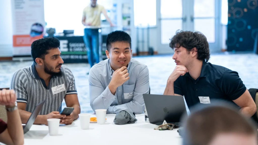 Three men seated at a table engaged in discussion, with laptops, cups, and name tags visible.