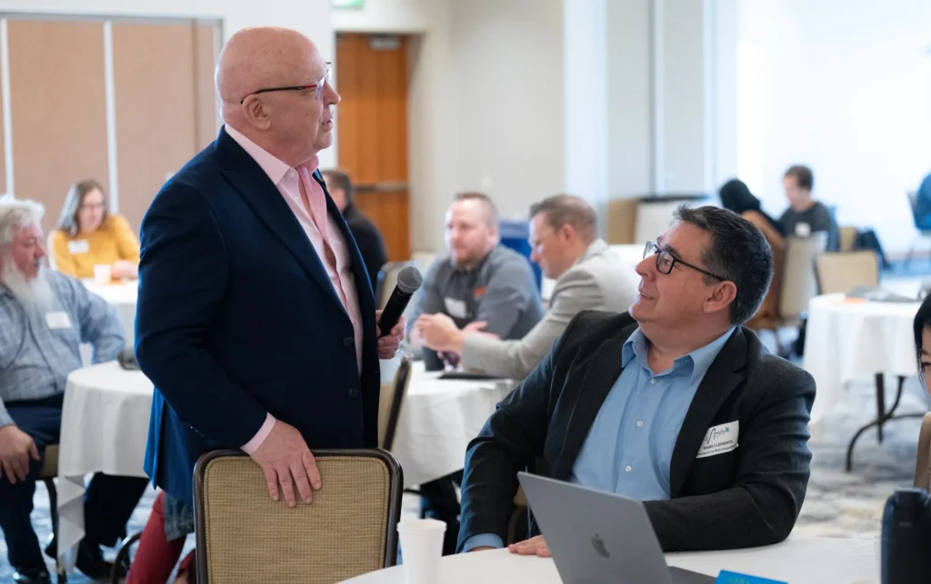 Two men in business attire conversing during a professional event with seated attendees.