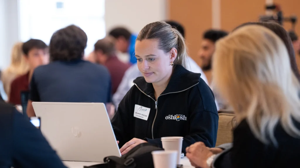 Young woman in black Oshkosh jacket working on laptop in a busy indoor setting.