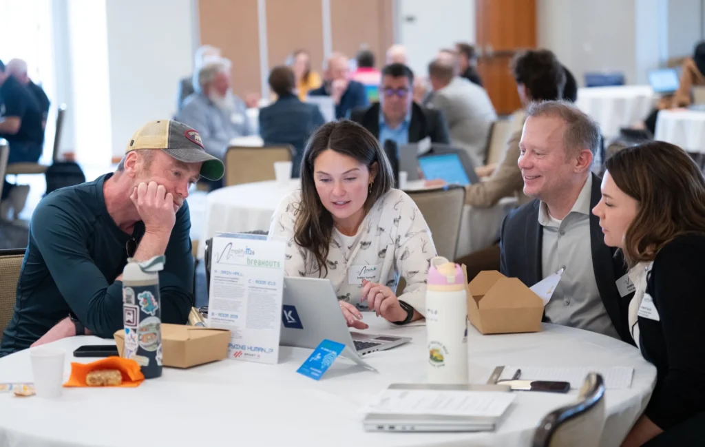 Group of four adults engaged in discussion around a table with laptops and documents in a conference setting.