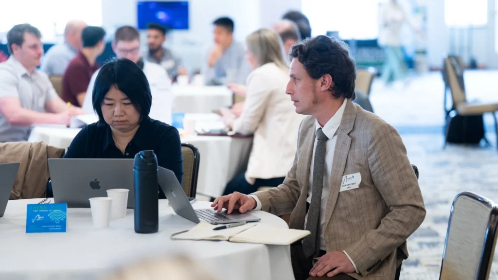 Two professionals working on laptops at a round table in a bright conference room.