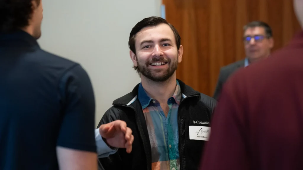 Smiling man wearing a black Columbia jacket and multicolored shirt, with name tag "Ari Thomas," engaging in conversation.