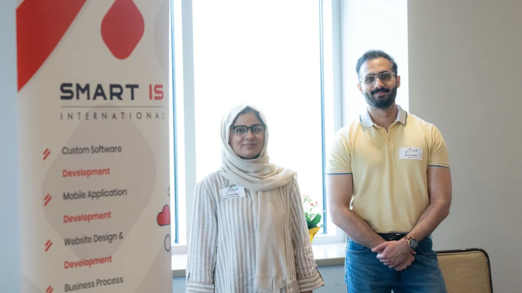 Two professionals standing beside a "SMART IS International" banner in a well-lit office.
